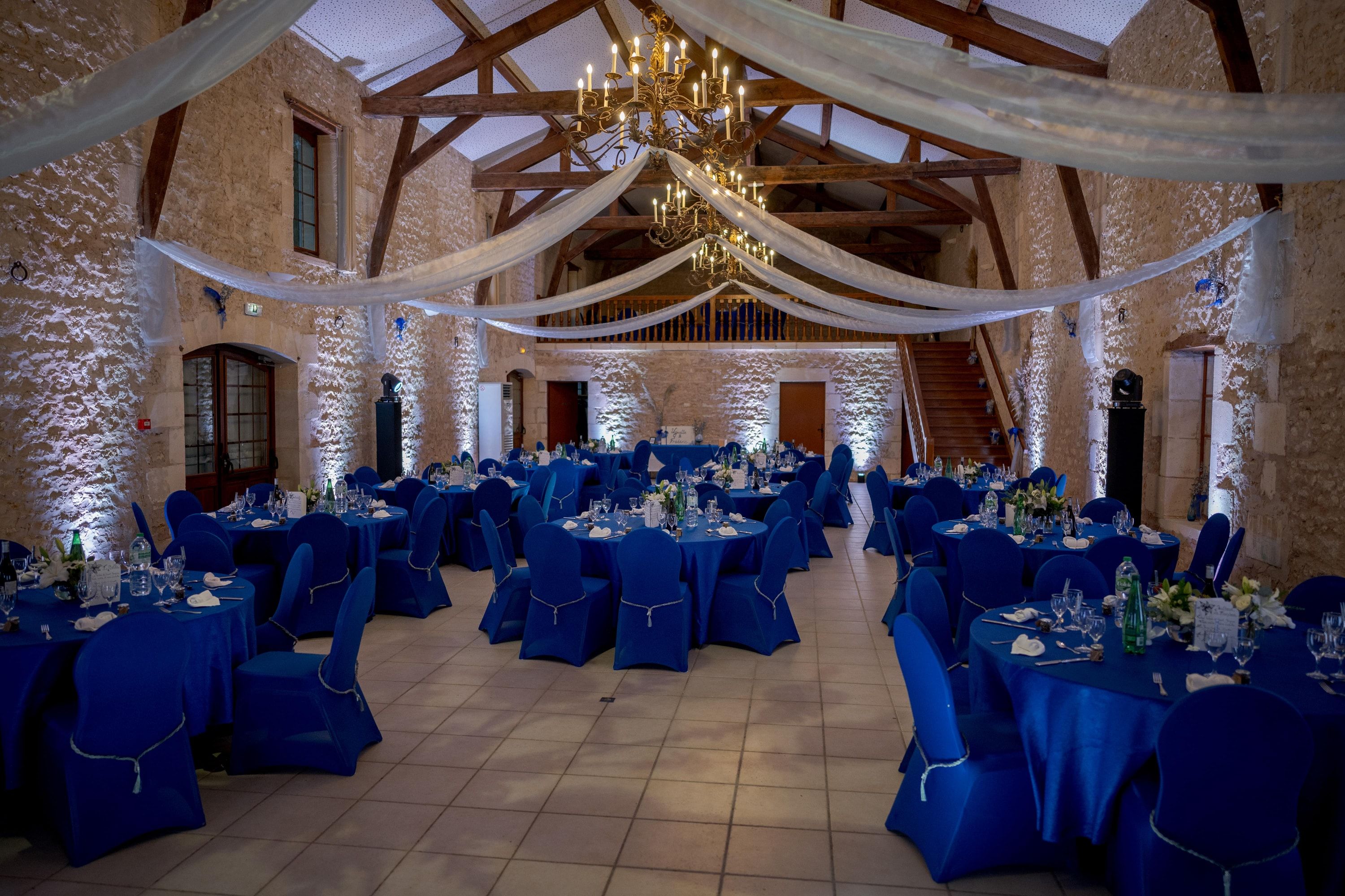 Salle de réception en pierre révélée par l’éclairage architectural, tables nappées de bleu